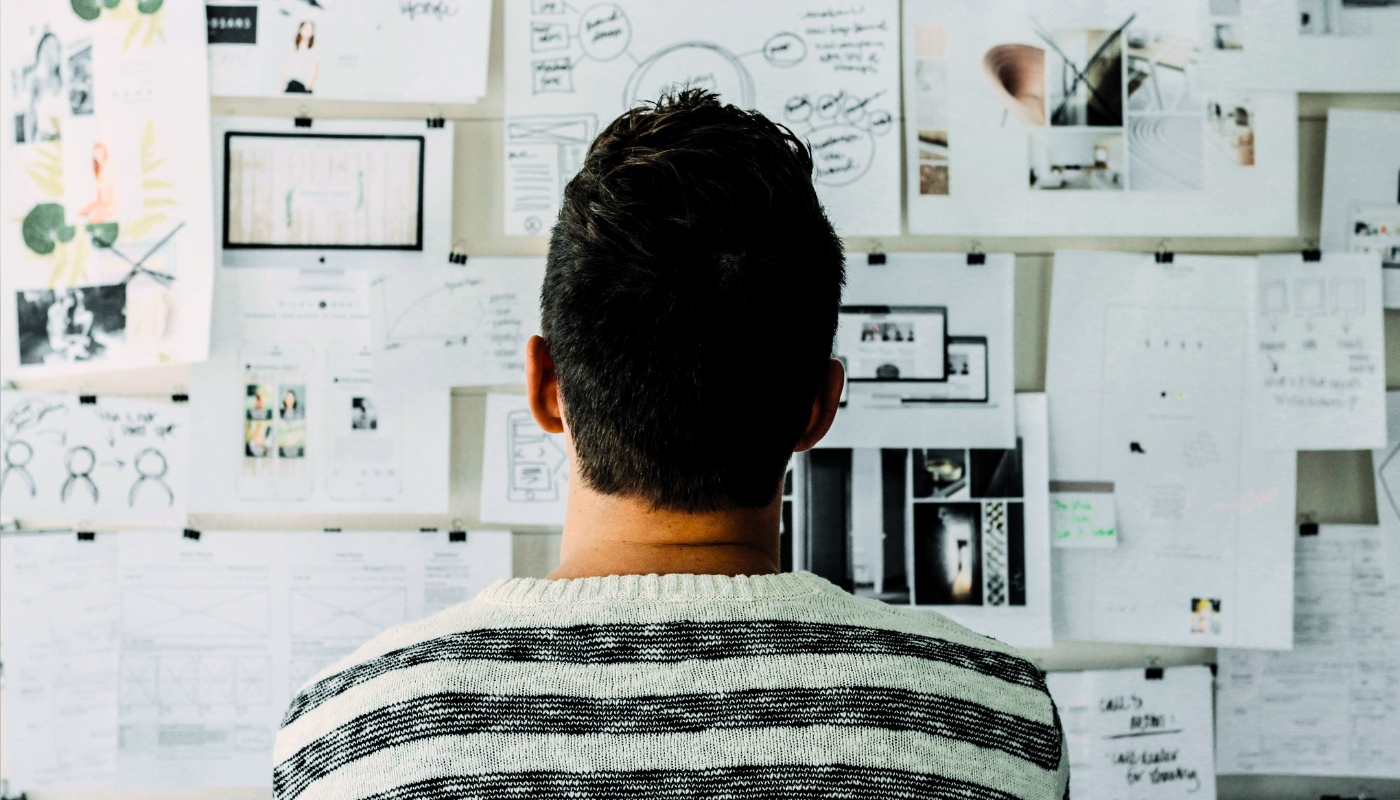 Person standing in front of a wall covered with notes, sketches, and planning documents, reviewing ideas and strategy concepts.